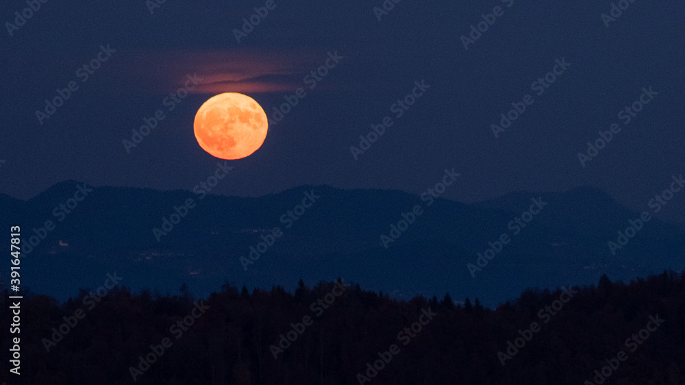 huge orange full moon rising behind a mountain ridge illuminating misty clouds creating a mystical atmosphere. glowing super moon rising into the night sky with layers of hills in the foreground.