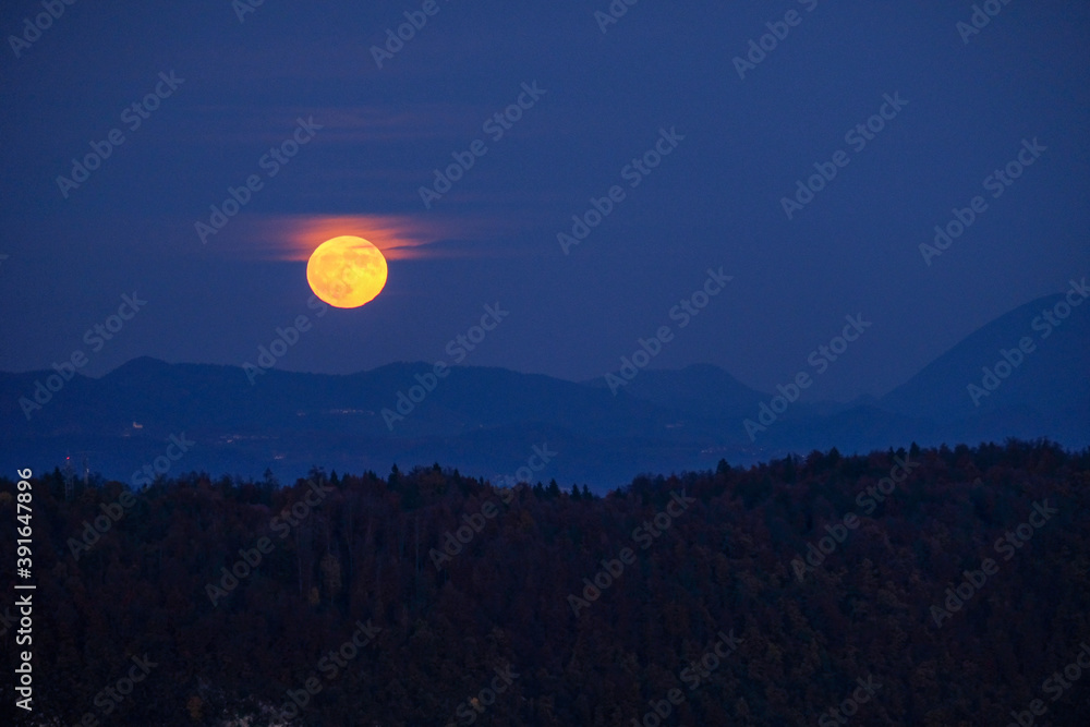 Fototapeta premium huge orange full moon rising behind a mountain ridge illuminating misty clouds creating a mystical atmosphere. glowing super moon rising into the night sky with layers of hills in the foreground.