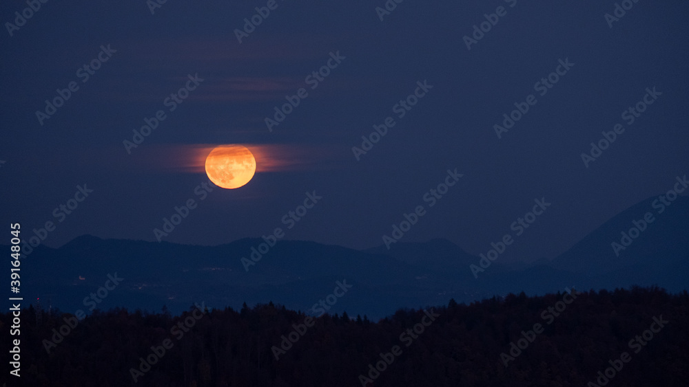 huge orange full moon rising behind a mountain ridge illuminating misty clouds creating a mystical atmosphere. glowing super moon rising into the night sky with layers of hills in the foreground. huge orange full moon rising behind a mountain ridge illuminating misty clouds creating a mystical atmosphere. glowing super moon rising into the night sky with layers of hills in the foreground.
