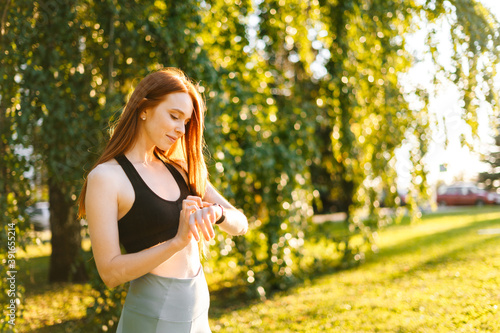 Portrait of fitness woman touch on the screen of the smart watch on her wrist to setting fitness app before running. Female at city park in sunny summer morning preparing sporty training,