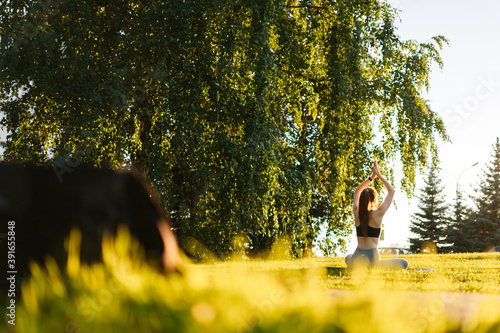 Back view of unrecognizable flexible young woman sitting on yoga mat in lotus position and raising hands up outside in city park. Remote view of female practicing yoga outdoors in sunny day. © dikushin