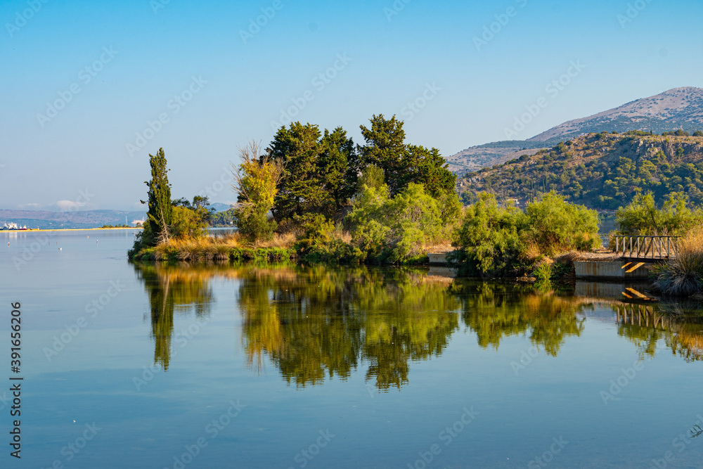 Fototapeta premium Koutavos Lagoon's serene waters mirror the charming town of Argostoli under a vast blue sky. Lush greenery frames the tranquil scene, offering a peaceful escape in the heart of Kefalonia, Greece