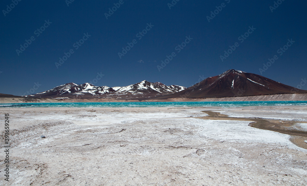 Alpine landscape in the cordillera. Beautiful turquoise glacier water lake in the Andes mountain range, surrounded by volcanoes, natural salt flats and dark mountains, under a deep blue sky.