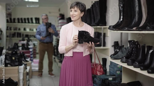 Beautiful senior woman choosing shoes in store and calling foe shop assistant as blurred man examining boots at the background. Customers making choice in shopping mall.