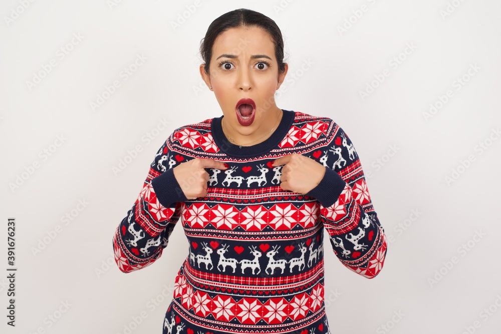 Embarrassed Young beautiful Arab woman wearing Christmas sweater, indicates at herself with puzzled expression, being shocked to be chosen to participate in competition, hesitates about something