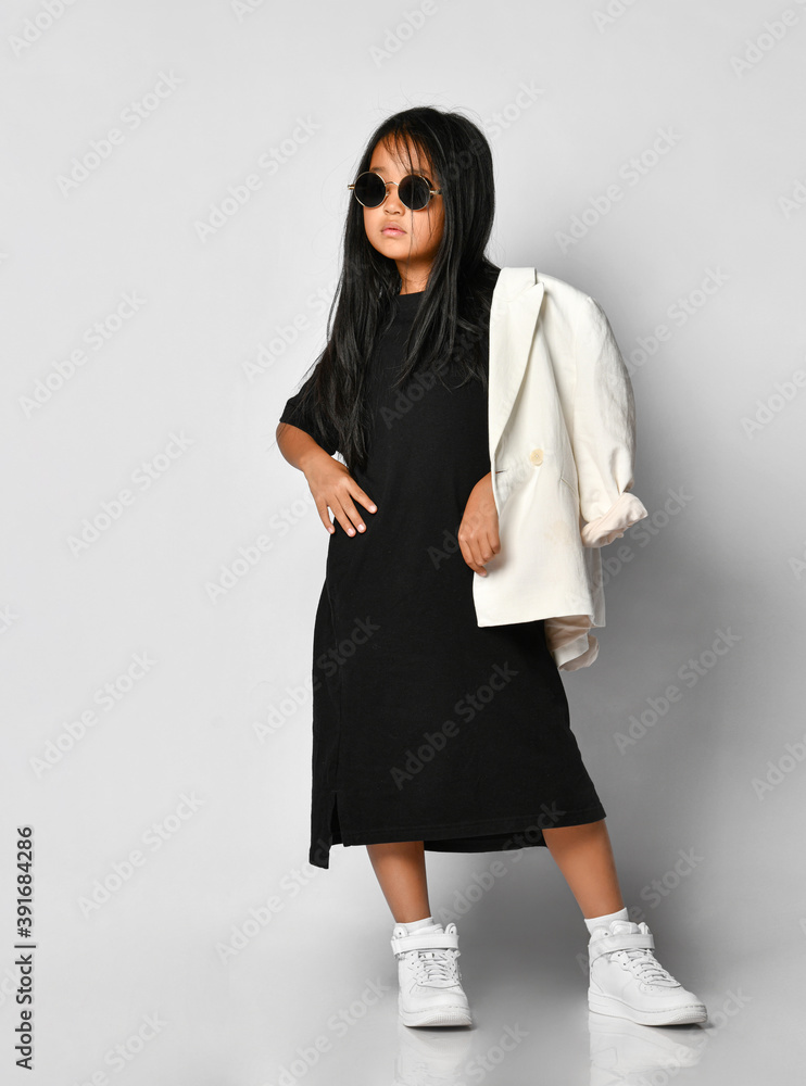 Fashionable portrait of a little Korean girl model posing professionally in the studio. Girl is wearing a black dress, white jacket, round sunglasses and white sneakers. Kids fashion concept.
