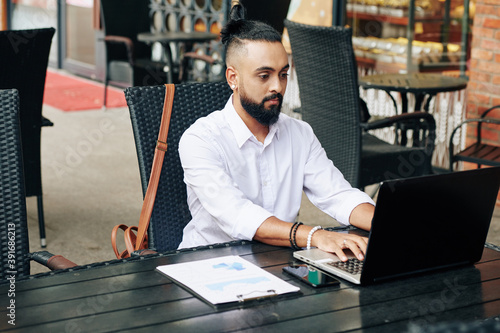 Serious young stylish businessman sitting at cafe table and working on laptop