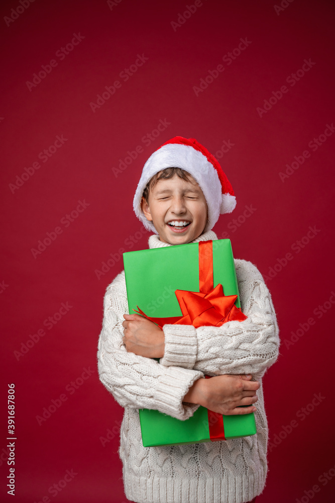 happy boy in a Santa hat holds a Christmas gift and closes his eyes happy boy in a Santa hat holds a Christmas gift and closes his eyes