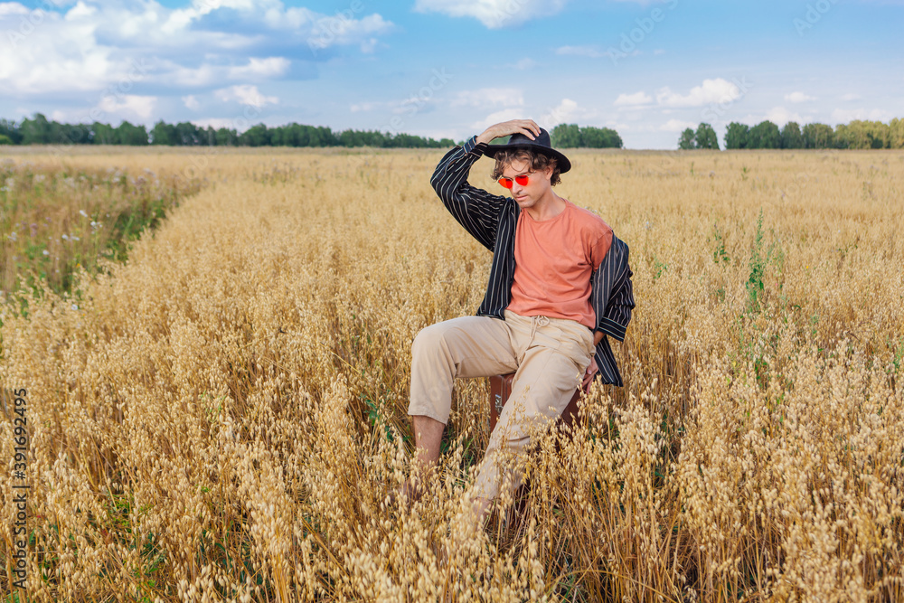 Tall handsome man with black hat and pink sunglasses sitting on a brown vintage leather suitcase at golden oat field. Tall handsome man with black hat and pink sunglasses sitting on a brown vintage leather suitcase at golden oat field.