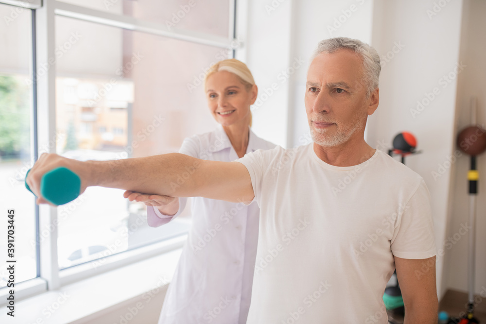 Blonde smiling therapist assisting a male patient during workout Blonde smiling therapist assisting a male patient during workout