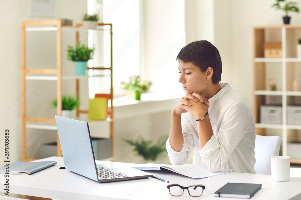 Pensive woman sitting at desk with laptop and notebook watching educational video or reading ebook Pensive woman sitting at desk with laptop and notebook watching educational video or reading ebook