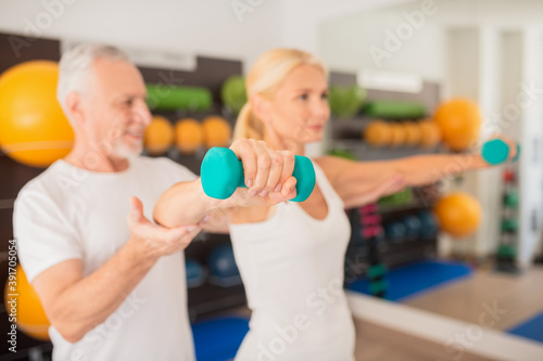 Male bearded instructor assisting blonde woman working with dumbbells in gym