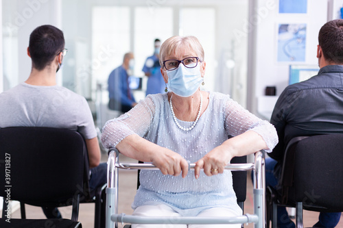 Disabled senior woman with face mask against coronavirus and walking frame looking at camera in hospital waiting area. Nurse assisting doctor during consultation in examination room.