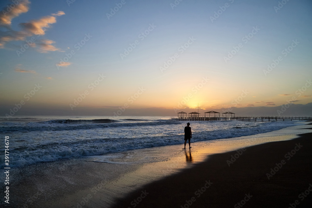View from beach to water of sea, waves with white foam and black silhouette of person in a nice evening during sunset with yellow sun. The concept of a holiday on the sea or ocean in the South