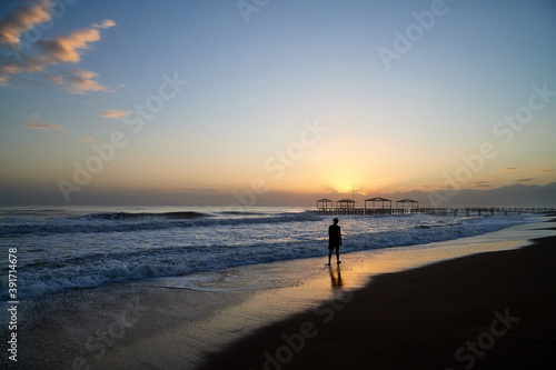 View from beach to water of sea, waves with white foam and black silhouette of person in a nice evening during sunset with yellow sun. The concept of a holiday on the sea or ocean in the South