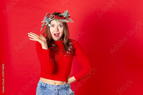 close up portrait of a surprised and dissatisfied young woman in a Christmas wreath, hands on hips, isolated on a red background.