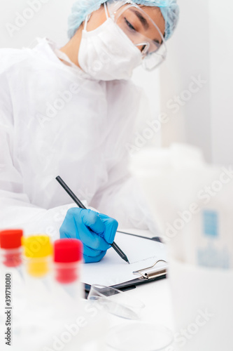 Medical worker in a protective suit fills out documents at the table in the medical office.Laboratory dishes on the table.Coronavirus and medical concept.