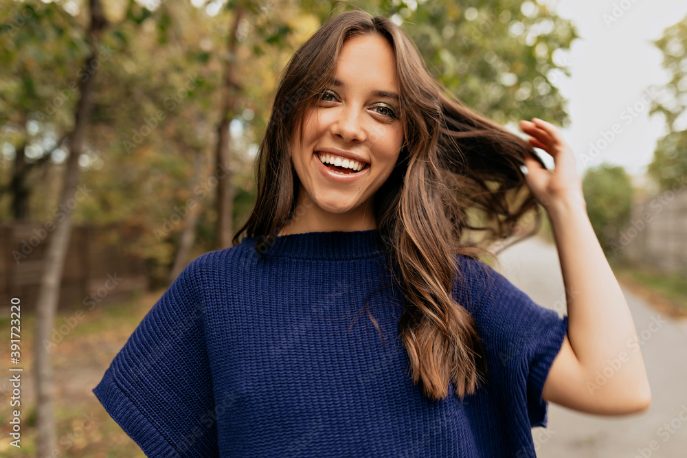 Happy smiling lady with wavy hair wearing blue sweater posing at camera and touching her hair in the park
