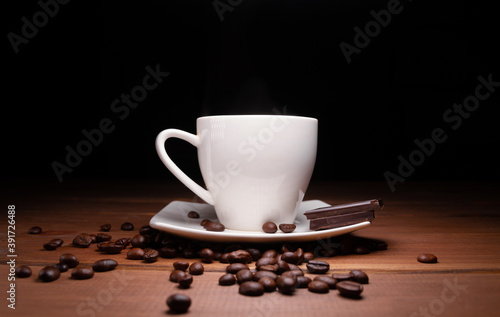 Small cup of hot coffee, pieces of delicious chocolate bar and fresh unground coffee beans scattered on a wooden table, with black background.