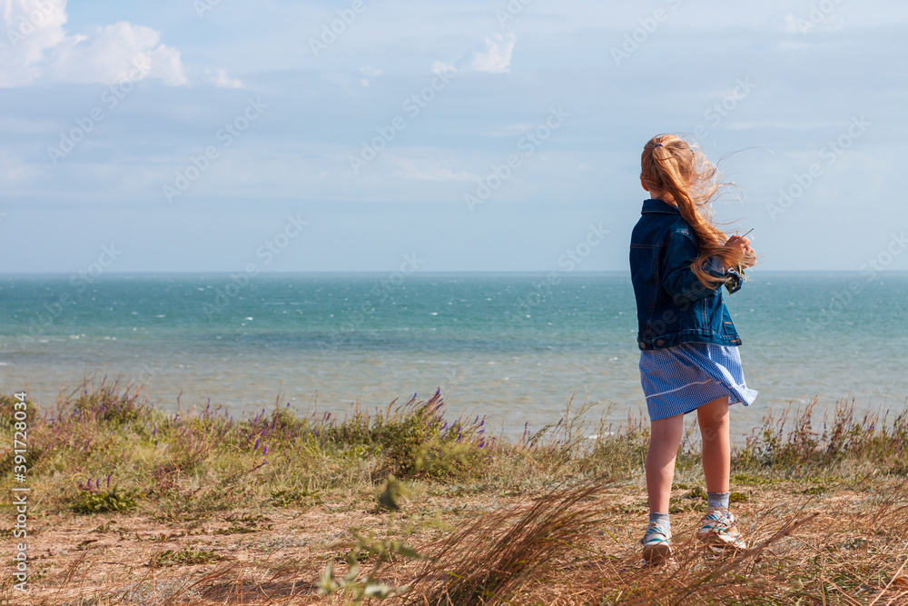 Adorable little girl in denim jacket back view on a hill with sea landscape view. Stylish child with long blonde hair on countryside cloudy sky background. Outdoor walking rural road trip faceless. Adorable little girl in denim jacket back view on a hill with sea landscape view. Stylish child with long blonde hair on countryside cloudy sky background. Outdoor walking rural road trip faceless.