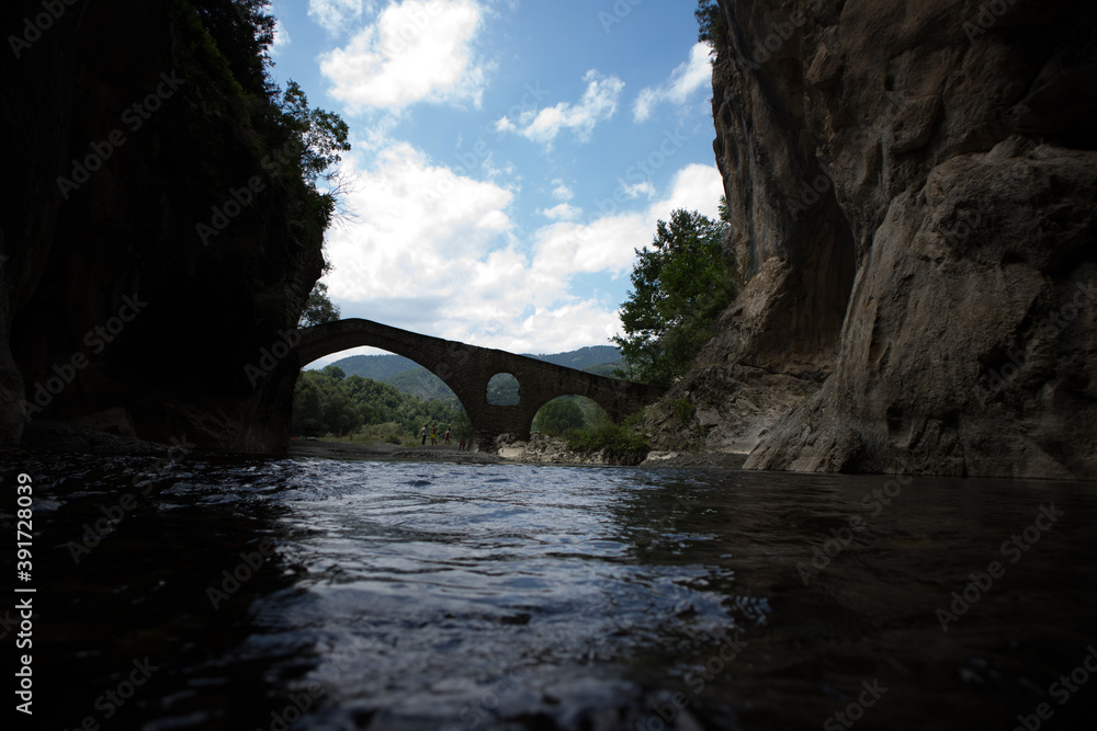 Fototapeta premium The beginning of a magnificent Gorge, with the silhouette of a stone bridge to appear at background and visitors near it. During the summer period, Portitsa, Grevena, Greece.