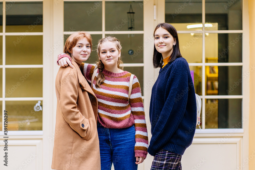 Two happy sisters embracing with their best friend in front of the school building outdoor. Sincere portrait of three female university students having fun on the way from school. Two happy sisters embracing with their best friend in front of the school building outdoor. Sincere portrait of three female university students having fun on the way from school.