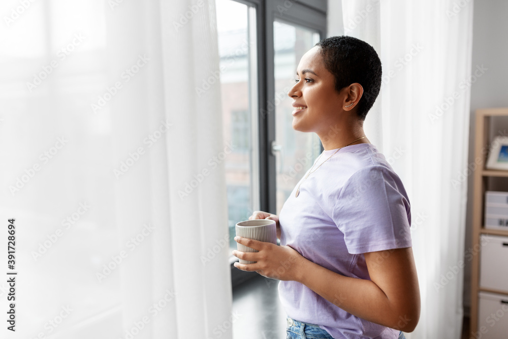 people and leisure concept - happy young african american woman with coffee cup looking out of window at home