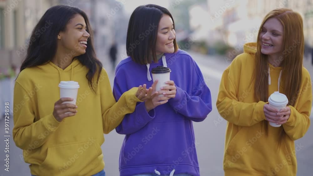 Multi-ethnic happy women walk together with a takeaway coffee in a recyclable cardboard cup outdoors. Young beautiful black, asian and caucasian female friends having fun in the city. Multi-ethnic happy women walk together with a takeaway coffee in a recyclable cardboard cup outdoors. Young beautiful black, asian and caucasian female friends having fun in the city.