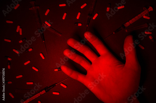 Drugs addiction concept. Drugs injection. Hand surrounded by used syringes and scattered capsules on black background under dim red light