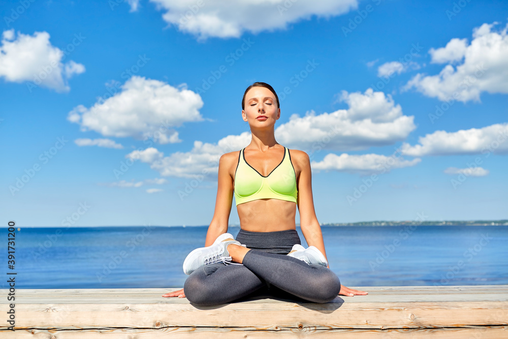 fitness, sport and yoga concept - young woman meditating in lotus pose at seaside