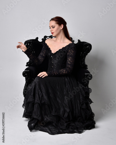 full length portrait of woman wearing black gothic dress, sitting on a ornate black armchair. Seated pose, against a studio background.