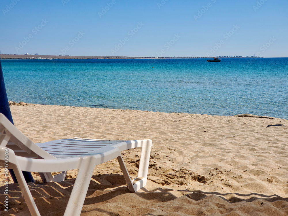 White, empty, plastic deck chair under the sun on the beach. Rest under the tropical sun. Sun loungers and parasols on the beach. Travel relax concept. Empty beach side.