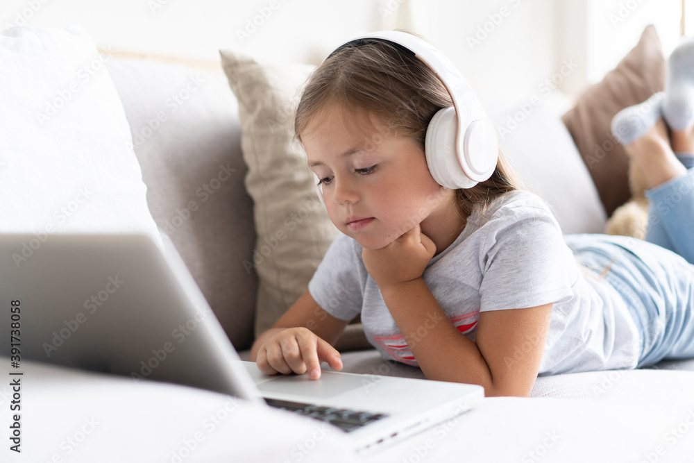 Online education of children. Sweet girl with headphones looking video lesson teacher conference laptop sitting on the couch at home. Online education of children. Sweet girl with headphones looking video lesson teacher conference laptop sitting on the couch at home.