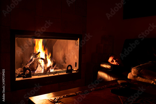 Teenager sleeping on the couch next to the fireplace with fire.