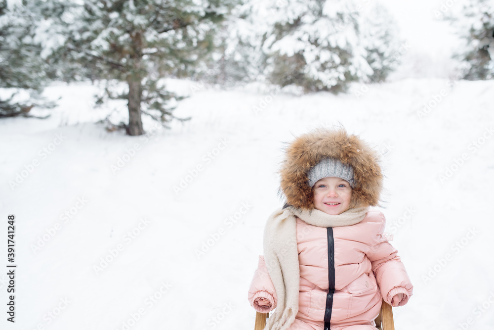 Fototapeta premium Little girl in a fur-trimmed hat laughs while sledding in winter 