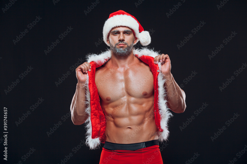 Fototapeta premium Sexy Santa Claus. Bodybuilder young handsome santa clause smile wearing Santa Claus hat shows off abs cubes at New Years eve and Christmas winter holiday black background.