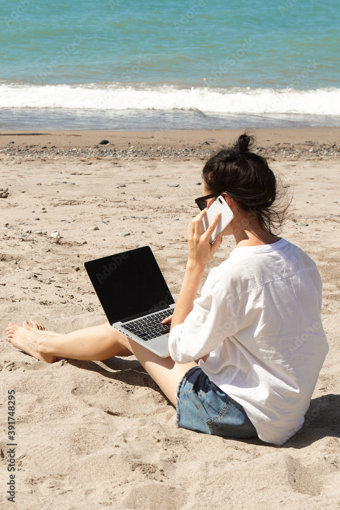 Young woman using laptop computer and talking on the phone on a beach. Vacation lifestyle communication. Freelance work concept