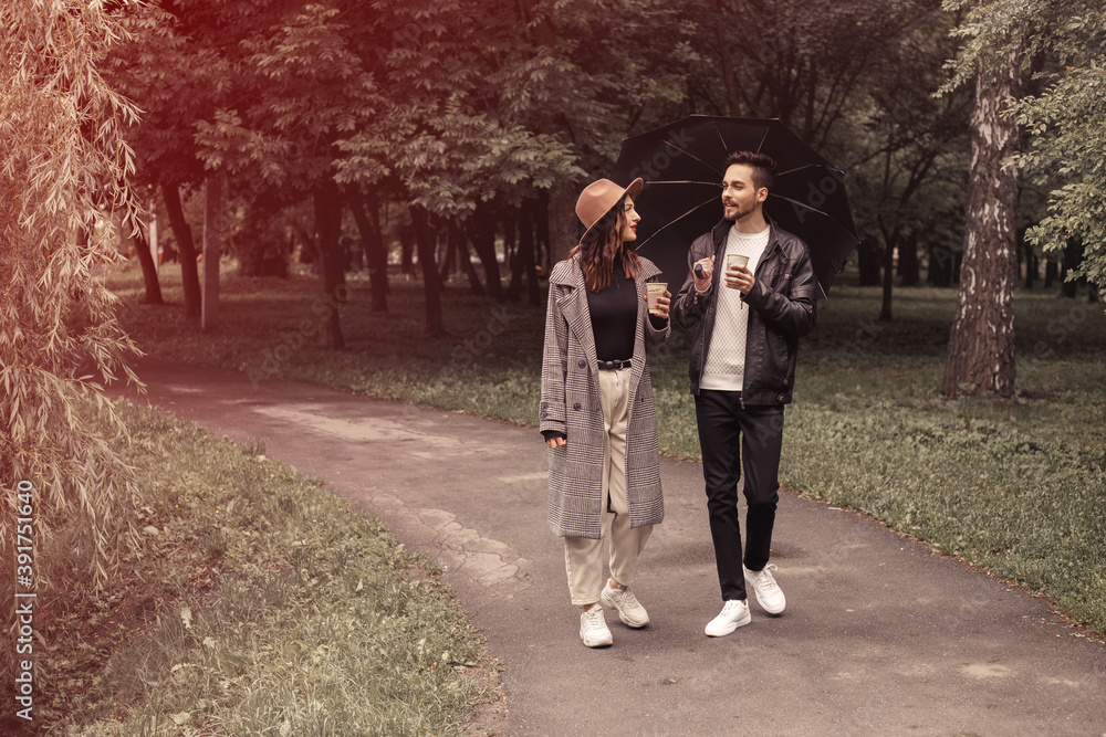 Young couple walking spring green city park, drinking hot coffee paper eco recycle cup. Two lovers husband wife spending time together, discuss news movie film black umbrella to shelter the rain. Young couple walking spring green city park, drinking hot coffee paper eco recycle cup. Two lovers husband wife spending time together, discuss news movie film black umbrella to shelter the rain.