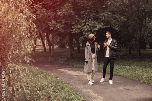 Young couple walking spring green city park, drinking hot coffee paper eco recycle cup. Two lovers husband wife spending time together, discuss news movie film black umbrella to shelter the rain.