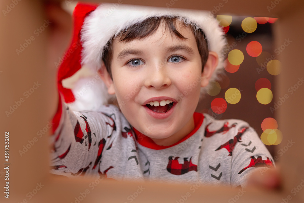 A surprised, happy, handsome European boy in a Santa Claus hat takes a present out of the box A surprised, happy, handsome European boy in a Santa Claus hat takes a present out of the box