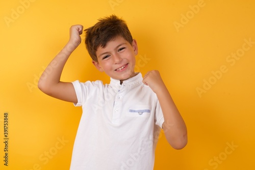 Attractive Young handsome Caucasian little boy standing against yellow wall celebrating a victory punching the air with his fists and a beaming toothy smile.