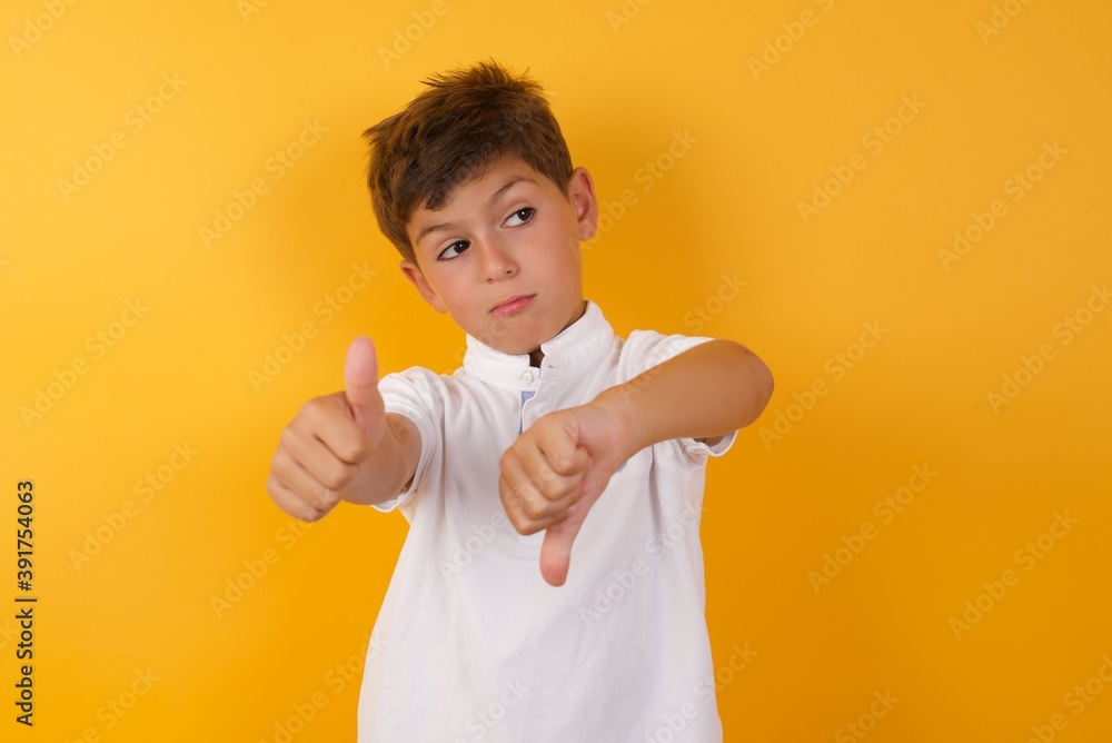 Young handsome Caucasian little boy standing against yellow wall feeling unsure making good bad sign. Displeased and unimpressed. Young handsome Caucasian little boy standing against yellow wall feeling unsure making good bad sign. Displeased and unimpressed.