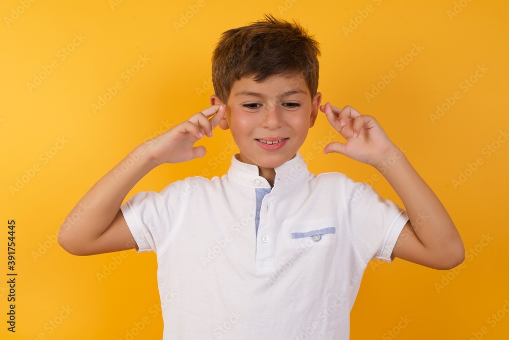 Young handsome Caucasian little boy standing against yellow wall holding fingers crossed with worried expression hoping boss didn't noticed mistakes at work. Young handsome Caucasian little boy standing against yellow wall holding fingers crossed with worried expression hoping boss didn't noticed mistakes at work.