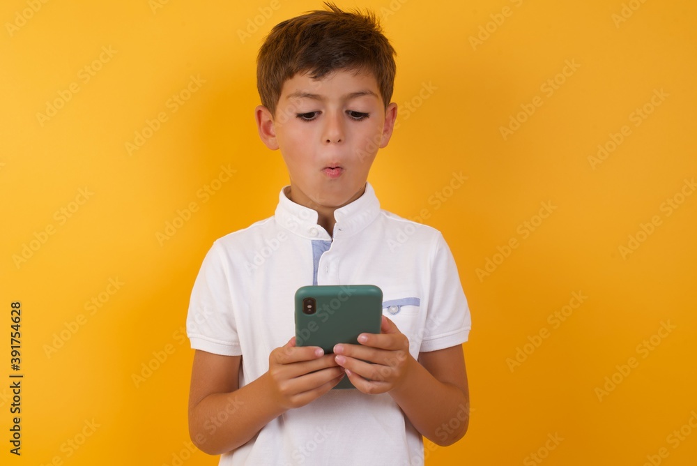 Young handsome Caucasian little boy standing against yellow background looks with bugged eyes, holds modern smart phone, receives unexpected message from friend, reads reminder. Young handsome Caucasian little boy standing against yellow background looks with bugged eyes, holds modern smart phone, receives unexpected message from friend, reads reminder.