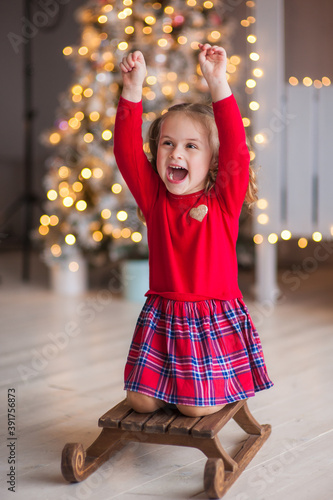 A cute little girl in a red dress sits on a wooden sled and joyfully raised her hands up in the Christmas interior with a Christmas tree. Christmas and New Year concept