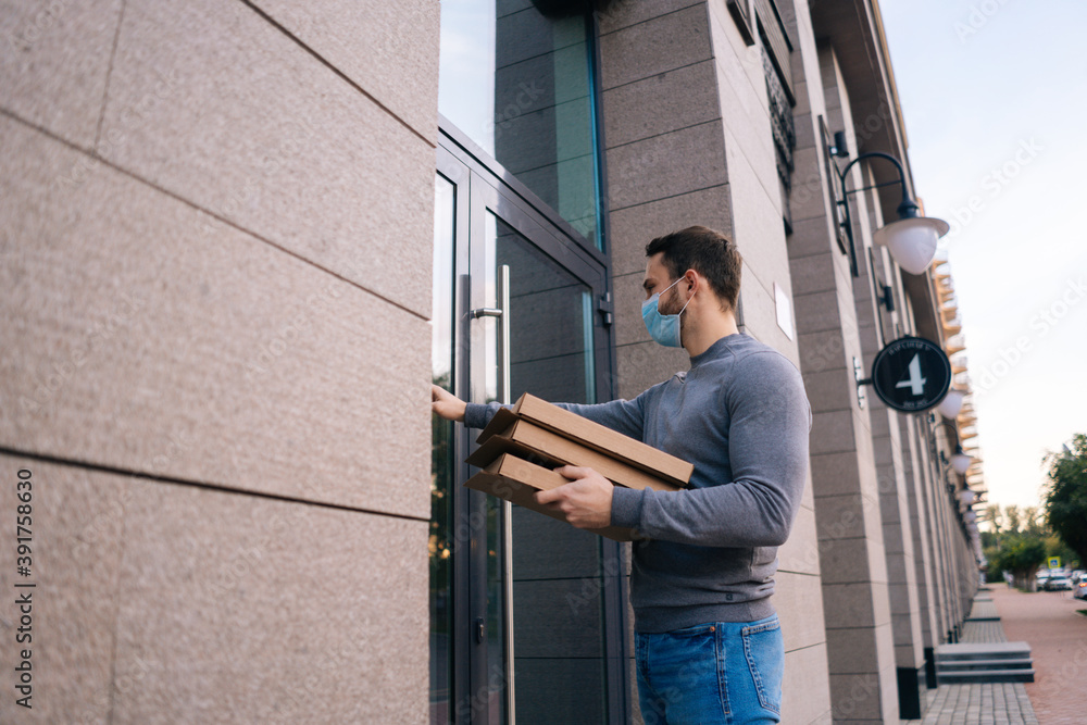 Delivery man wearing medical mask ringing door for delivery carton boxes with hot pizza. Deliveryman in protective mask holding box with food. Concept of food delivery service during quarantine. Delivery man wearing medical mask ringing door for delivery carton boxes with hot pizza. Deliveryman in protective mask holding box with food. Concept of food delivery service during quarantine.