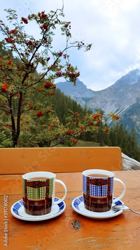 Two coffee mugs with traditional dirndl outfit painting on an outdoor table against the beautiful mountains of the Austrian Alps.