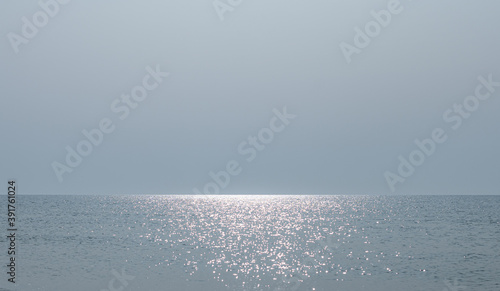 A calm sea with light ripples and sun glare in the center and a cloudness serene sky. A clear line of the horizon in the seascape, gray-blue tones.