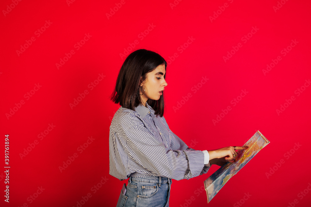 Beautiful impressed girl is looking and pointing with finger at paper map isolated on red background. Travel concept. Beautiful impressed girl is looking and pointing with finger at paper map isolated on red background. Travel concept.