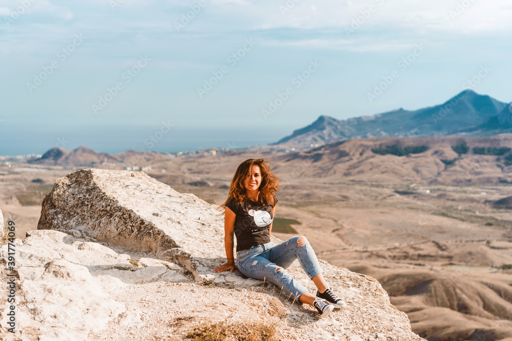 A young beautiful woman with long hair and jeans sits on a rock in the Koktebel valley in Crimea. The landscape of sandy hilly desert.